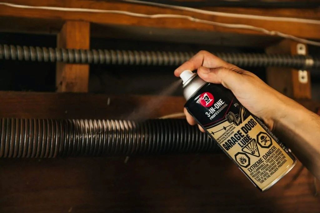 Professional technician lubricating garage door rollers and tracks using silicone-based spray lubricant